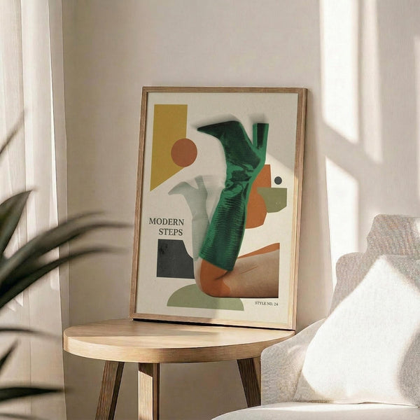 May include: Abstract geometric art print in wooden frame on round wood side table next to beige upholstered chair. Artwork features collaged elements including green high-heeled boot, yellow and orange shapes, brown circle, and text reading "Modern Steps" against cream background. Natural lighting casts soft shadows across white wall and plant leaves visible at left edge.