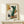 May include: Framed abstract art print in light wood frame leaning against white wall on wooden shelf. Artwork features geometric shapes in mustard yellow, burnt orange, sage green, and black with person wearing emerald green ribbed sweater and brown pants. Text reads "Modern Steps" and "Style No 24". Small wrapped white gift box with greenery sits on shelf in front of frame.