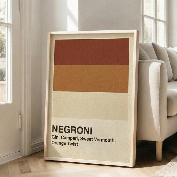 May include: Framed poster with horizontal color blocks in rust red, golden orange, and cream tones, displaying "NEGRONI" and cocktail ingredients text in black letters. Light wood frame leaning against white wall next to beige upholstered sofa in bright, naturally-lit living room with large windows.