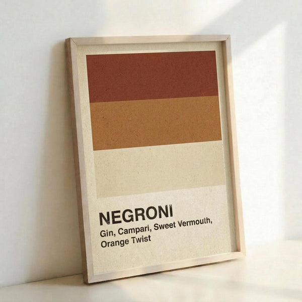 May include: Framed minimalist poster print leaning against white wall showing horizontal color blocks in burgundy, orange, tan and cream representing Negroni cocktail ingredients. Black text at bottom reads "NEGRONI Gin, Campari, Sweet Vermouth, Orange Twist" on cream background. Light wood frame with natural finish.