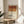 May include: Framed poster displaying a Negroni cocktail recipe with geometric color blocks in rust red and golden orange tones against cream background. The print shows ingredients list including gin, Campari, sweet vermouth, and orange twist in black text. Wooden frame sits on round wooden side table next to beige upholstered chair with plant leaves visible, all in bright natural sunlight creating soft shadows on white walls.