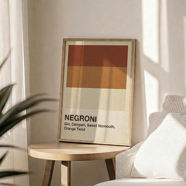 May include: Framed poster displaying a Negroni cocktail recipe with geometric color blocks in rust red and golden orange tones against cream background. The print shows ingredients list including gin, Campari, sweet vermouth, and orange twist in black text. Wooden frame sits on round wooden side table next to beige upholstered chair with plant leaves visible, all in bright natural sunlight creating soft shadows on white walls.