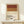 May include: Framed minimalist cocktail poster with horizontal color bands in rust red, tan, and cream representing Negroni ingredients, displayed on light wood console table against white wall. Small wrapped gift box with evergreen sprig sits beside the natural wood frame.