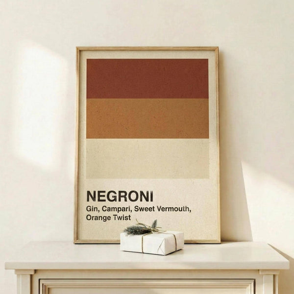 May include: Framed minimalist cocktail poster with horizontal color bands in rust red, tan, and cream representing Negroni ingredients, displayed on light wood console table against white wall. Small wrapped gift box with evergreen sprig sits beside the natural wood frame.