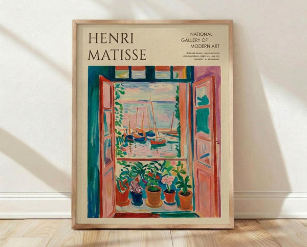 May include: Framed Henri Matisse exhibition poster featuring colorful Fauve-style painting of open French doors revealing boats on water, with potted plants on windowsill. Vibrant blues, oranges, pinks and greens in expressive brushstrokes. Light wood frame against white wall on wooden floor with natural lighting creating soft shadows.