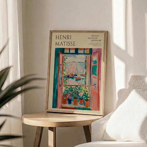 May include: Framed Henri Matisse poster displaying colorful fauve-style painting of open window with boats visible outside and potted plants on windowsill, featuring vibrant blues, oranges, and greens. Poster sits on round wooden side table next to beige upholstered chair, with plant leaves casting shadows on white wall in bright natural lighting.
