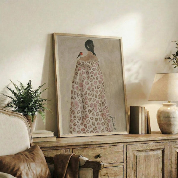May include: Framed artwork showing a woman with dark hair in a bun wearing a leopard print dress, with a small red bird perched on her shoulder, displayed on a rustic wooden sideboard alongside potted plants, books, and ceramic vases in a warm, neutral-toned living room setting.