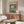 May include: Henri Matisse museum poster featuring colorful open window scene with boats and potted plants, displayed above wooden sideboard in modern living room with white upholstered chair, floor lamp, and glass coffee table on light wooden floors
