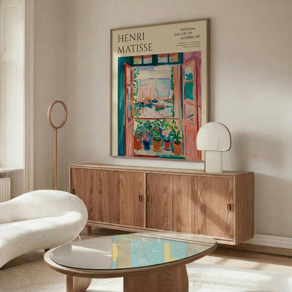May include: Henri Matisse museum poster featuring colorful open window scene with boats and potted plants, displayed above wooden sideboard in modern living room with white upholstered chair, floor lamp, and glass coffee table on light wooden floors