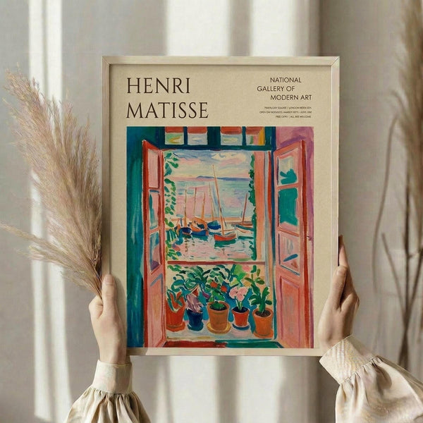 May include: hands holding a framed Henri Matisse exhibition poster featuring a vibrant Fauvist painting of an open window overlooking a harbor with boats, potted plants on the windowsill, rendered in bold oranges, greens, and blues. The cream-colored poster shows "National Gallery of Modern Art" text. Person wears a beige ruffled sleeve top while holding dried pampas grass, photographed against white curtains in soft natural lighting.
