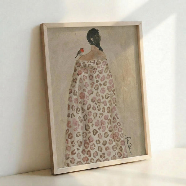 May include: Framed artwork leaning against white wall showing woman from behind wearing leopard print dress or robe in muted pink and brown tones, with black braided hair and small red bird perched on her shoulder, painted in soft watercolor style on beige textured background within light wood frame