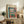May include: Framed Henri Matisse museum poster displayed on rustic wooden sideboard in modern living room. Poster shows colorful Fauvist painting of open window with view of boats on water, potted plants on windowsill, vibrant blues, reds, and greens. Natural wood frame complements surrounding decor including fern plant, ceramic vase, books, and neutral upholstered furniture.