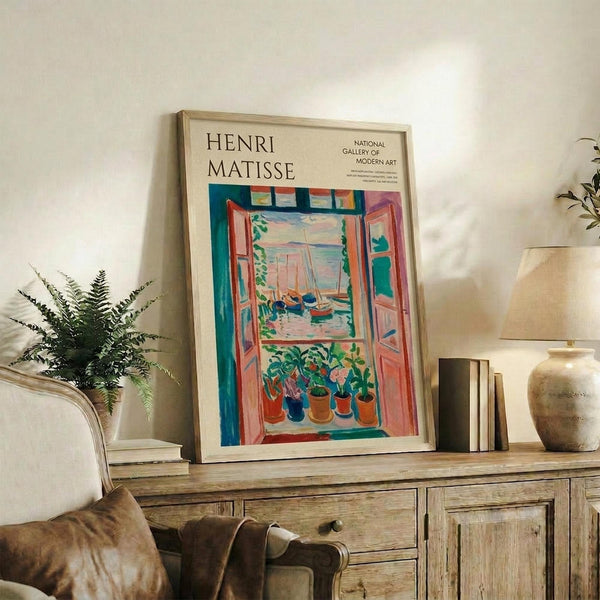 May include: Framed Henri Matisse museum poster displayed on rustic wooden sideboard in modern living room. Poster shows colorful Fauvist painting of open window with view of boats on water, potted plants on windowsill, vibrant blues, reds, and greens. Natural wood frame complements surrounding decor including fern plant, ceramic vase, books, and neutral upholstered furniture.