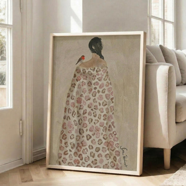 May include: Framed artwork leaning against white wall next to beige sofa in bright room with large windows. The print shows a woman from behind wearing a leopard print blanket or shawl with a small red robin perched on her shoulder. Her dark hair is in a braid against the neutral beige background. The piece has a light wood frame and soft, watercolor-like style with muted earth tones.