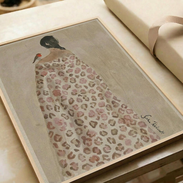 May include: Framed art print displayed on white bedding showing a figure in leopard print dress with black hair and small red bird perched on shoulder. The artwork features muted pink and brown leopard spots on cream background with artist signature visible. Print appears in light wood frame against soft white linens and pillows.
