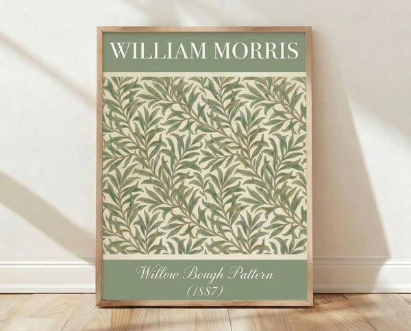 May include: Framed poster with sage green header reading "William Morris" above an intricate botanical pattern featuring flowing willow branches and leaves in muted green tones on cream background. Bottom green banner shows "Willow Bough Pattern (1887)" in white script. Natural wood frame leans against white wall on light wooden floor with soft shadows.