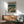 May include: Framed anime-style poster showing figure in trench coat standing in futuristic cityscape with glowing signs and tall buildings, displayed above wooden sideboard with white ceramic lamp in modern living room with mint green mid-century armchair and white walls with natural sunlight shadows.