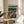 May include: Framed anime-style poster featuring a character in tan trench coat standing in rainy cyberpunk cityscape with teal and orange tones, displayed on round wooden side table next to beige upholstered chair with white pillow, plant visible in corner, natural lighting creates soft shadows on white wall