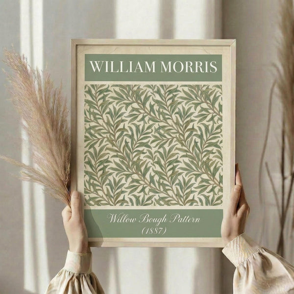 May include: Hands holding a framed botanical art print featuring William Morris's 1887 Willow Bough pattern in sage green tones on cream background. The print shows flowing willow branches and leaves in an intricate repeating design. Light wood frame with white matting against neutral curtained window. Person wearing beige ruffled sleeves holds dried pampas grass stems, creating a soft, natural styling scene.