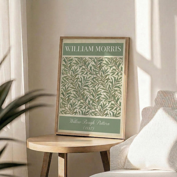 May include: Framed botanical art print leaning on round wooden side table next to beige upholstered chair. The poster features green willow branch pattern on cream background with sage green header and footer text bands. Natural wood frame catches soft window light creating shadows on neutral wall. Small potted plant visible in corner of serene, minimalist room setting.