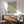 May include: Framed William Morris botanical print featuring green willow branch pattern on cream background, displayed above wood sideboard with table lamp. Mid-century modern living room with sage green upholstered chair, round coffee table, white walls with dramatic sunlight shadows creating geometric patterns across the space.