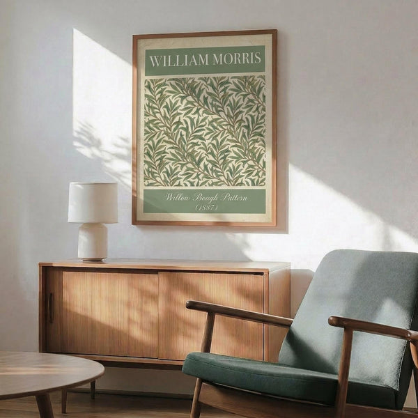 May include: Framed William Morris botanical print featuring green willow branch pattern on cream background, displayed above wood sideboard with table lamp. Mid-century modern living room with sage green upholstered chair, round coffee table, white walls with dramatic sunlight shadows creating geometric patterns across the space.