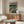 May include: Vintage-style anime poster mounted on white wall above wooden sideboard, featuring character in trench coat walking through neon-lit cyberpunk cityscape with Japanese text header. Modern living room setting with cream upholstered chair, glass-top coffee table, and minimalist white table lamp. Warm wood tones complement teal and orange poster colors.