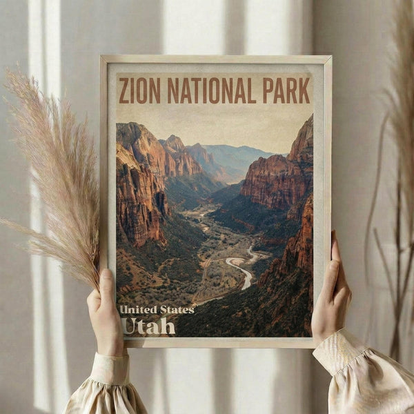 May include: Person holding framed vintage-style travel poster featuring Zion canyon landscape with towering red rock cliffs, winding river, and desert valley. Warm earth tones dominate the design. Light wood frame against neutral curtained background with dried pampas grass visible on left side.