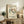 May include: Framed vintage exhibition poster featuring five dancing figures in blue, brown, and green tones, displayed on rustic wooden sideboard alongside fern plant, ceramic vase, and stacked books in warm-lit living room setting with cream upholstered furniture.