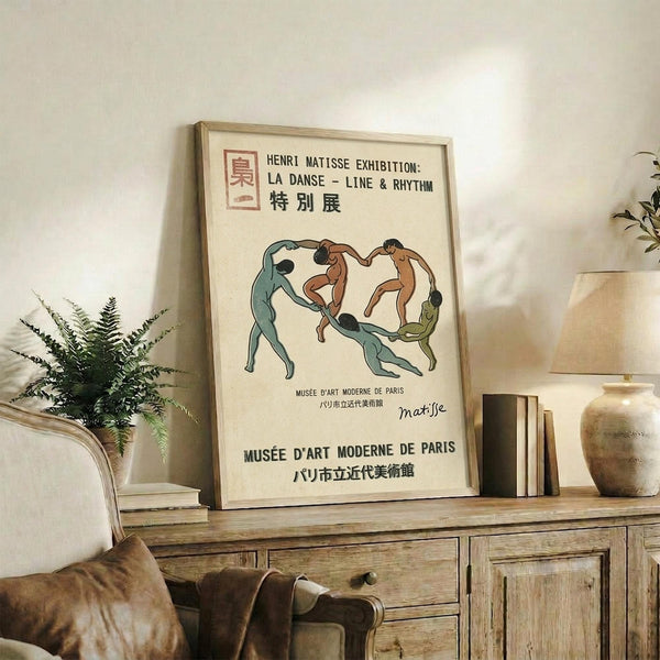 May include: Framed vintage exhibition poster featuring five dancing figures in blue, brown, and green tones, displayed on rustic wooden sideboard alongside fern plant, ceramic vase, and stacked books in warm-lit living room setting with cream upholstered furniture.