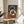 May include: Abstract artwork with three interlocking rings in terracotta orange, cream, and dark brown on a teal and dusty rose background, displayed in a thin wooden frame on a round wooden side table next to a beige upholstered chair, with natural sunlight casting shadows and plant leaves visible in the corner of a neutral-toned room.