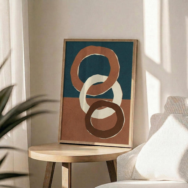May include: Abstract artwork with three interlocking rings in terracotta orange, cream, and dark brown on a teal and dusty rose background, displayed in a thin wooden frame on a round wooden side table next to a beige upholstered chair, with natural sunlight casting shadows and plant leaves visible in the corner of a neutral-toned room.