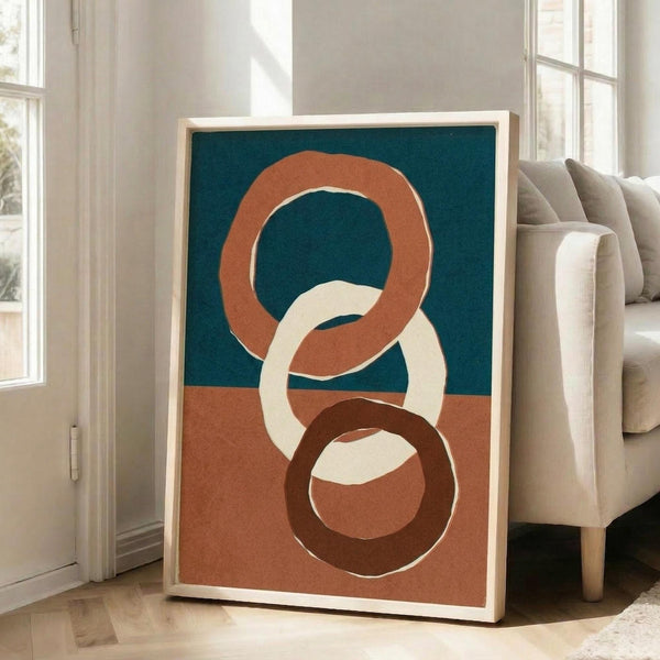 May include: A framed abstract art print leaning against a beige sofa in a bright living room with white walls and large windows. The artwork features three interlocking circular rings in terracotta orange, cream, and dark brown on a split background of teal blue and coral pink. The print has a light wood frame and casts a subtle shadow on the hardwood floor.