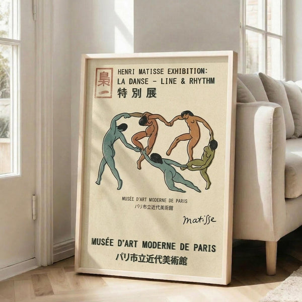 May include: Framed exhibition poster featuring five dancing figures in a circular formation rendered in muted teal, brown, and olive green tones against cream background. Japanese and French text indicates Henri Matisse exhibition at Musée d'Art Moderne de Paris. Light wood frame leans against white wall next to beige upholstered sofa in bright, minimalist living room with large windows and natural light casting shadows.