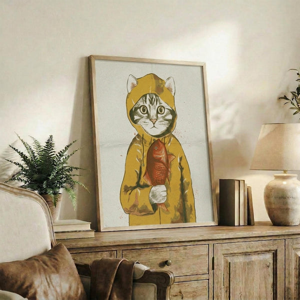 May include: Framed artwork featuring a tabby cat wearing a yellow hooded raincoat and holding an orange fish, displayed on a rustic wooden sideboard alongside books, ceramic vases, a fern plant, and cream-colored furniture in a warmly lit living room setting.