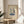 May include: Framed minimalist line art print showing two people dancing on top of a turntable record player, rendered in blue tones on cream background. Print sits on round wooden side table next to beige upholstered chair with throw pillow. Natural sunlight streams across neutral wall with plant leaves visible in corner.