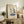 May include: Framed minimalist poster showing two dancing figures in blue tones emerging from a turntable record player, displayed on rustic wooden sideboard alongside fern plant, ceramic vase, and stacked books in warm neutral living room setting with cream armchair visible