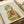 Framed print showing a tabby cat wearing a yellow hooded raincoat and holding a red fish. The artwork has a vintage illustration style with muted colors on light background. The wooden frame sits on a white surface next to cream-colored ribbon, creating a soft, neutral setting.