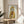 May include: Framed artwork showing a tabby cat wearing a bright yellow hooded raincoat and holding an orange fish, displayed on a round wooden side table next to a beige upholstered chair. Natural sunlight streams across the neutral-toned room, with a plant visible in the corner. The illustration has a whimsical, detailed style in a thin natural wood frame.