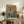 May include: Framed poster with Japanese text and illustration of astronaut figure and green mecha robot against orange sky with futuristic city skyline, displayed on rustic wooden sideboard alongside fern plant, ceramic vase, and books in cozy living room setting with beige armchair