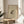 May include: Framed vintage Japanese-style ink drawing of a spotted frog on cream paper with Chinese characters and red seal stamp, displayed on round wooden side table next to beige upholstered chair with white pillow, green plant leaves visible, natural sunlight creating soft shadows on white wall