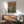 May include: Framed vintage-style anime poster featuring a mech robot and pilot character in green space suits against a futuristic cityscape, displayed above a wooden credenza with white ceramic lamp in a modern living room with mint green mid-century armchair and natural lighting casting shadows on white walls.