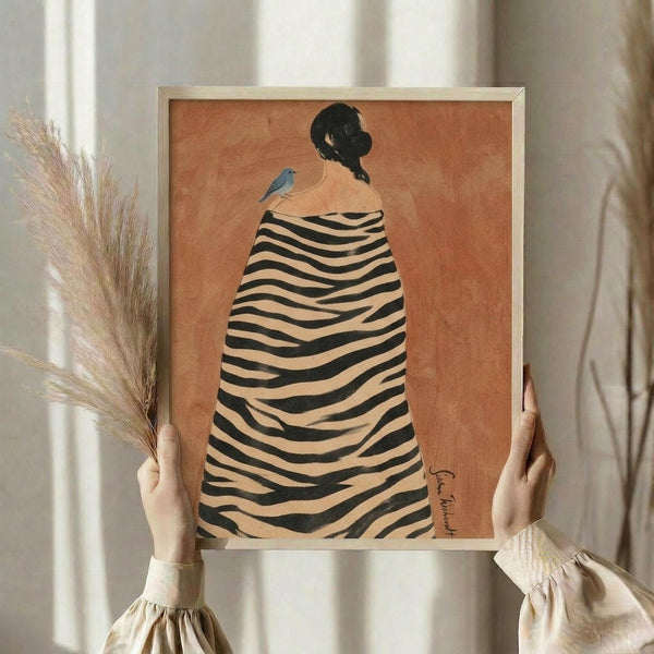May include: Hands holding a framed art print showing a woman from behind wearing a black and cream zebra-striped cloak with a small blue bird perched on her shoulder, set against a warm orange background. The artwork is displayed in a light wooden frame, with dried pampas grass visible to the left and soft natural lighting from nearby windows.