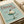 May include: Vintage-style poster with mint green background featuring elegant cocktail glass filled with white whipped cream topped with red cherry. Red serif text reads "Dinner Club" at top, "The art of indulgence" on left, "A dash of decadence" on right. Print displayed in thin light wood frame on white bedding with cream ribbon visible at edge.