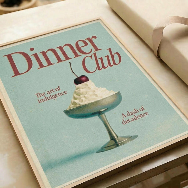 May include: Vintage-style poster with mint green background featuring elegant cocktail glass filled with white whipped cream topped with red cherry. Red serif text reads "Dinner Club" at top, "The art of indulgence" on left, "A dash of decadence" on right. Print displayed in thin light wood frame on white bedding with cream ribbon visible at edge.