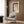 May include: Abstract art print with terracotta pink and cream watercolor blocks, black brushstroke, and vertical dots, displayed in white frame on wood-paneled wall above beige upholstered chair with geometric pillow, alongside brass floor lamp and dark side table with open book