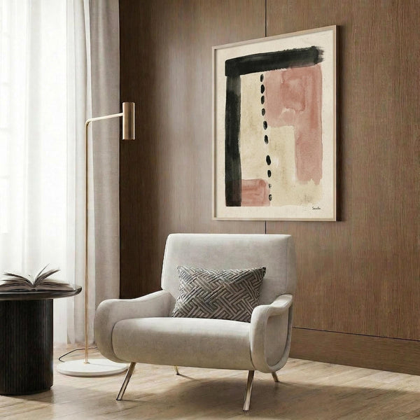 May include: Abstract art print with terracotta pink and cream watercolor blocks, black brushstroke, and vertical dots, displayed in white frame on wood-paneled wall above beige upholstered chair with geometric pillow, alongside brass floor lamp and dark side table with open book