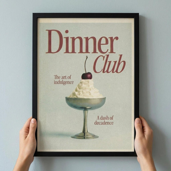 May include: Hands holding a black-framed vintage-style poster featuring red text "Dinner Club" and "The art of indulgence" with an illustration of whipped cream dessert topped with a cherry in a glass coupe, plus "A dash of decadence" text, displayed against a light gray wall in a cream and muted green color palette.