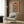 May include: Modern living room with beige upholstered chair and geometric pillow, brass floor lamp, dark wood paneled wall, and framed artwork showing a figure in black and white striped zebra-print cloak against warm orange background, displayed in light wood frame above seating area.