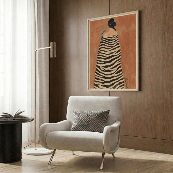 May include: Modern living room with beige upholstered chair and geometric pillow, brass floor lamp, dark wood paneled wall, and framed artwork showing a figure in black and white striped zebra-print cloak against warm orange background, displayed in light wood frame above seating area.
